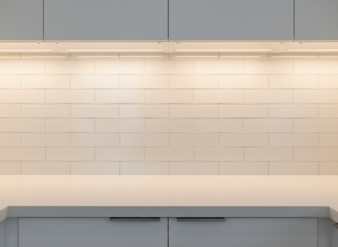 A detailed shot of a freshly tiled kitchen backsplash in a renovated Toronto apartment, featuring elongated subway tiles in a soft matte white with very thin, evenly spaced light gray grout lines. Under-cabinet LED strip lighting runs along the edge of the upper cabinets, washing the tiles with warm light and creating subtle shadows that emphasize the precise alignment. The bottom of sleek white cabinetry and the top of a light quartz countertop frame the backsplash. Photographic realism, captured straight-on at eye level, with sharp focus and a shallow depth of field that slightly softens the outer edges. The mood is clean, modern, and meticulous, showcasing the tilework quality that a professional general contractor and renovation specialist provides.